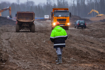 Ust-Luga, Leningrad oblast, Russia - November 16, 2021: Worker go on dirty road. Heavy construction machines on background, blurred. © Alexey Rezvykh