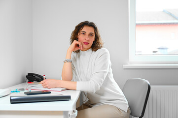 A confident and sophisticated woman with curly hair wears a creamy white sweater in her immaculately organized office. Her professional appearance and expertise demonstrate her leadership in the field