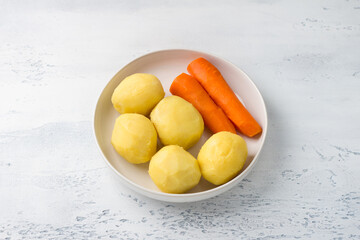 White plate with peeled boiled vegetables, potatoes and carrots on a light blue background, top view. Vegetable salad cooking step.