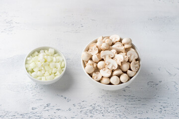 White bowls with chopped mini champignons and onions on a light blue background, top view. Stage of cooking a healthy vegan dish