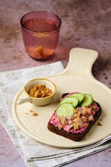 Smorrebrod on rye bread with beetroot mousse, pickled herring, fresh cucumber slices and crispy onion on a ceramic board on a brown background.