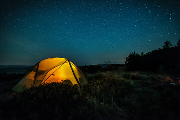 Glowing camping tent in the mountain forest under a starry sky