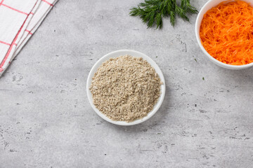 Bowls with ground sunflower seeds, carrots and a bunch of fresh dill on a gray textured background, top view. Stage of cooking delicious vegan cutlets or other vegetable dish