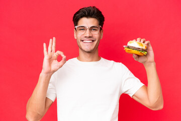 Young Caucasian man holding a burger isolated on red background showing ok sign with fingers