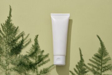 Minimal scene of a white cosmetic container placed on a light background with several green leaves....