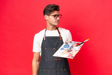 Young artist caucasian man holding a palette isolated on red background looking to the side