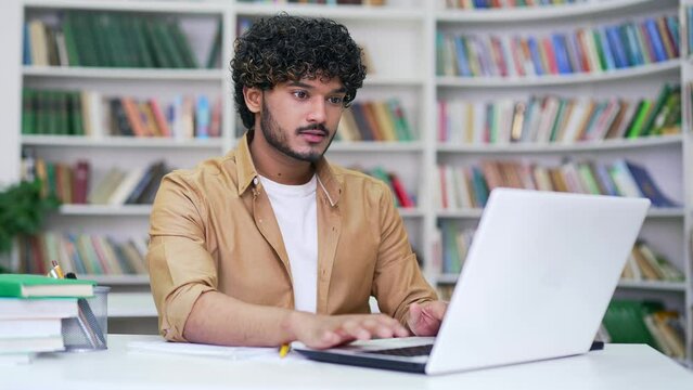 Young student on online e-learning taking notes or video calling remote teacher using laptop computer in campus library space. Handsome male applicant is preparing for university exams in classroom