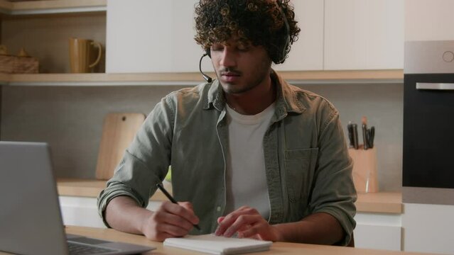 Distant e-learning home education Arabian man in headset watching online computer class laptop webinar at kitchen Indian guy student internet teacher studying writing notes cram SAT test prep course