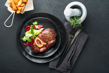 Steaks Osso Buco. Sliced beef grilled Osso Buco or porterhouse meat steak with garnished with salad and french fries on black marble board on old wooden background. Top view. Mock up.
