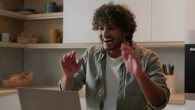 Happy Indian man smiling talking casual video call with laptop at kitchen Arabian guy talk to computer online virtual conference greeting hello chat showing ok share news internet conversation at home