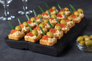 Festive canapes with red caviar, cheese and olives on croutons of white bread on dark board on gray background