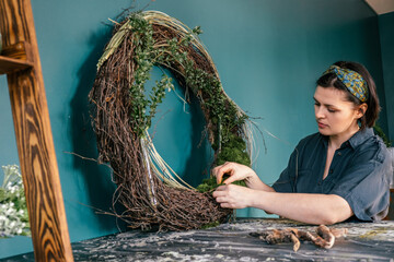 Attractive young woman makes home decor from natural materials in the workshop.The process of making a wreath of twigs,stabilized moss.and leaves.Biophilia design concept.