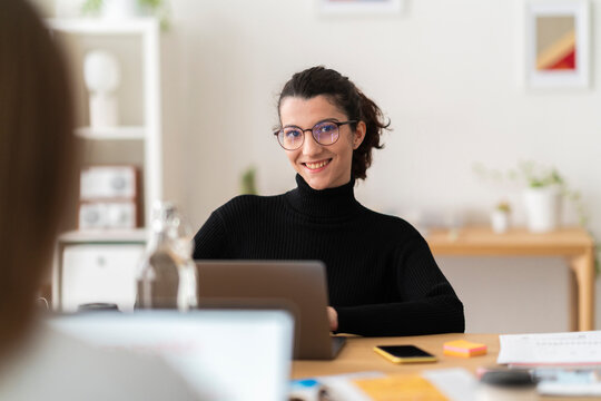 Positive young female in casual clothes smiling and looking at camera while sitting at table working on project at computer in bright modern location