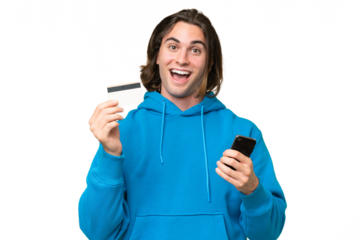 Young handsome man isolated on green chroma background buying with the mobile and holding a credit card with surprised expression