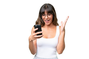 Fototapeta premium Young caucasian woman over isolated background looking at the camera while using the mobile with surprised expression