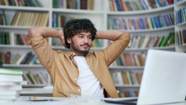 Happy young university student finished homework on laptop computer sitting in campus library space. Satisfied smiling college teacher relaxes, pauses, stretches on chair with hands behind head