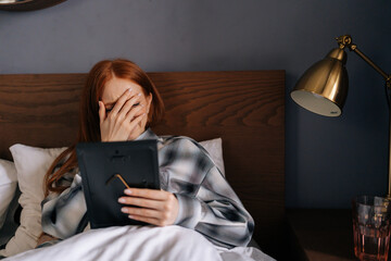 Portrait of grief-stricken young woman lying on bed covering face with hand and crying while holding picture frame, touching photograph with love. Concept of nostalgia, grief, longing and loneliness.