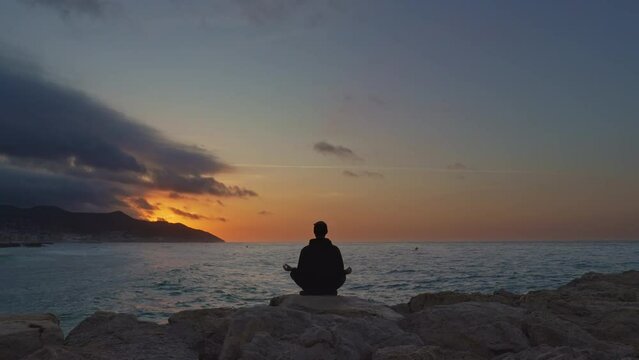 Timelapse captures zen man sitting in lotus posture and meditating on rocks as golden sun rises above sea