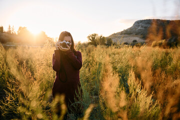 A young woman strolling through the countryside with her vintage camera at sunset.