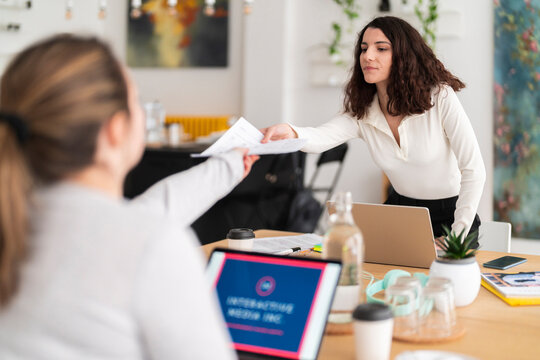 Positive young female in casual clothes sitting at table with laptop and passing around a sheet documents while working on project in creative workspace