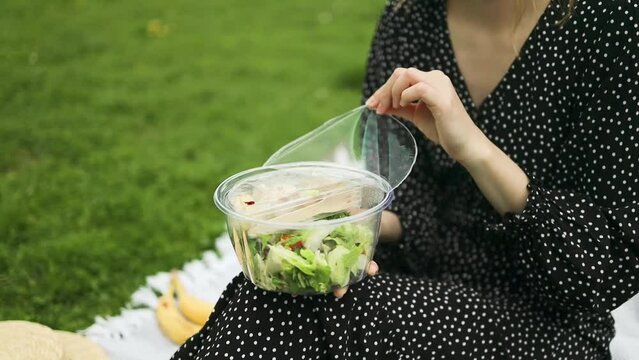 Caucasian woman hands opens plastic wrap with fresh salad sitting at a picnic in the park in the city. High quality FullHD footage