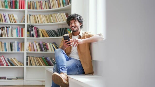 Young smiling university student using smartphone while sitting on bench in campus library space. Happy male is chatting online with friends, browsing social networks, checking messages or texting