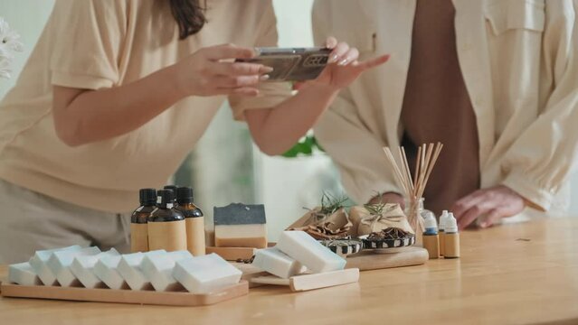 Cropped shot of two unrecognizable women taking photos of beautifully packed organic cosmetic products on smartphone while working in small beauty and skincare shop