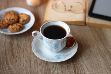 Cup of tea or coffee, plate of cookies, books, e-reader, pencil and lit candles on the table. Selective focus.