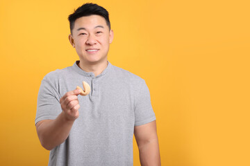 Asian man holding tasty fortune cookie with prediction on orange background. Space for text