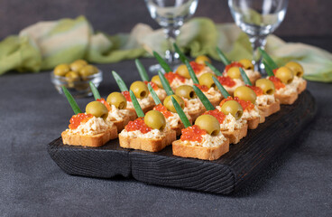 Festive canapes with red caviar and olives on croutons of white bread on dark board on gray background