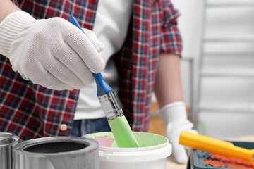 Man dipping brush into bucket of green paint indoors, closeup