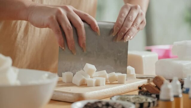 Cropped shot of unrecognizable woman preparing premade soap base for melting by dicing it on wooden cutting board in workshop
