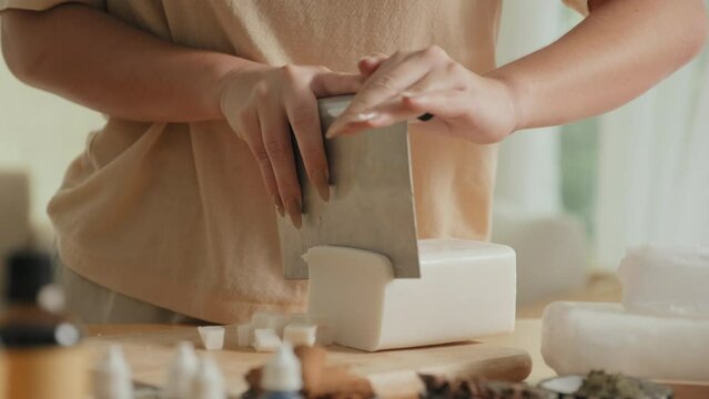 Tilt down shot of young Caucasian woman slicing premade soap base on cutting board while making organic cosmetics in bright workshop