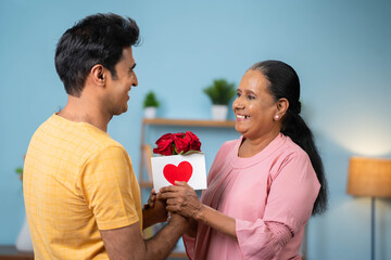 Happy Indian son wishing Mother day by giving greeting card with flowers to elderly mother at home - concept of relationship, bonding and enjoyment