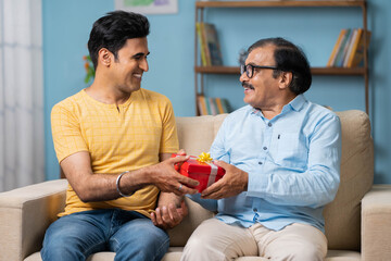Happy smiling indian adult son giving surprise gift or present to father while sitting on sofa at...