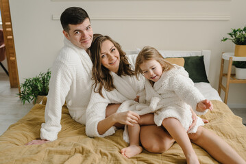 Happy mom, dad hugging girl kid smiling. Young mother, father hugs cute little daughter in bathrobes lying on bed, look at camera in hotel room. Healthcare concept. Family with child having fun.