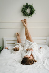 Young mother with cute little daughter in bathrobe lying, putting their feet, legs up on bed....