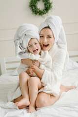 Young mother with cute little daughter in bathrobe, towels on heads sitting lovely on bed. Healthcare and family concept. Cheerful girls having beauty day at home. Happy mom hugging kid. Closeup.