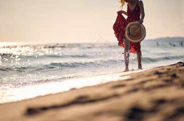 Young woman in waving red summer dress walking on beach holding fashionable straw hat. Vacation, holiday, lifestyle concept.
