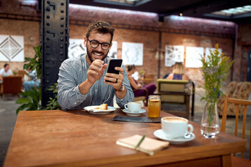 Young cheerful man at cafe smiling and looking at phone. Enjoying cup of coffee and cake.
