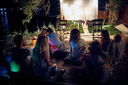 A group of people sitting on the floor in the bar on the river bank and enjoying night cinema. Night, summer, bar, river