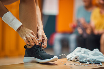 Young woman tying her shoe in gym locker room, with earphones and towel on bench, getting ready for workout.