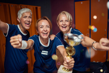 Team of two senior and one young women in dressing room, holding golden cup and feeling victorious.