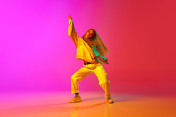 Fototapeta na wymiar Portrait with one charming girl, dancer with dreadlocks wearing hip-hop style of clothes and dancing over gradient pink background in neon light