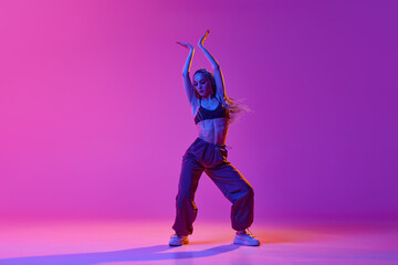 Young beautiful slim girl, dancer wearing hip-hop clothes dancing over gradient purple neon...