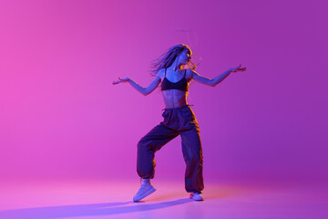 Fototapeta na wymiar One young professional hip-hop dancer wearing stylish clothes dancing with inspiration over purple background in neon light. Beauty of movement