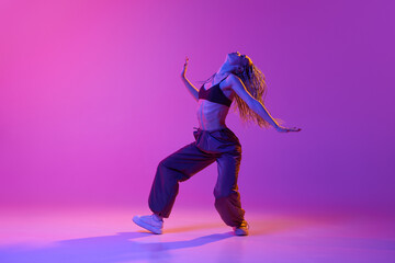 Fototapeta na wymiar One attractive young woman, girl with pigtails dancing solo performance with pleasure over purple studio background in neon light. Freestyle