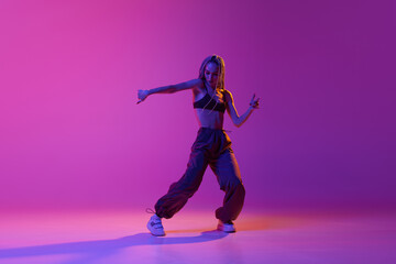 Fototapeta na wymiar Portrait with modern young dancer wearing fashion clothes in motion over puple background in neon light. Liberty of dance