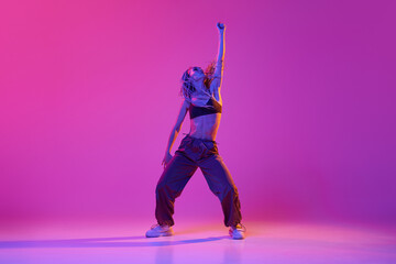 Young beautiful slim girl, modern dancer wearing hip-hop clothes dancing over gradient purple neon background. Contemporary dance style - obrazy, fototapety, plakaty