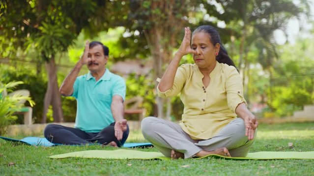 Indian senior couple doing pranayama or nostril breathing yoga during morning at park - concept of mindfulness, relationship and healthy retirement lifestyle